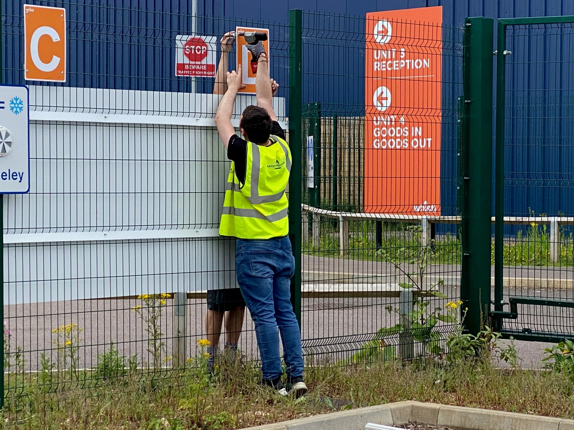 Sign fitter installing orange goods out and unit reception signs at a Whistl gate